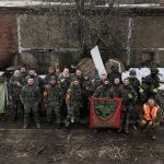 Группа участников в защитной экипировке позирует со знамёнами у заснеженного здания.