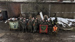 Группа участников в защитной экипировке позирует со знамёнами у заснеженного здания.