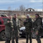 Четверо участников в защитной экипировке стоят у автомобилей на полигоне.