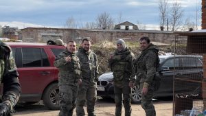 Четверо участников в защитной экипировке стоят у автомобилей на полигоне.