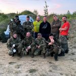 Участники ночной игры в защитной экипировке фотографируются у походного лагеря.